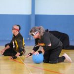 três pessoas praticam goalball. À frente, uma mulher segura a bola junto ao chão