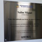 Placa em homenagem a Salim Miguel com os dizeres - "Salim Miguel (1924-2016). Uma vida dedicada aos livros, às artes e à cultura. Diretor executivo da Editora da YUFSC de 1983 a 1991. Título Doutor Honoris Causa pela UFSC em 2002. Homenagem da Gestão Universidade Presente ao centenário de Salim Miguel em 30/01/2024.