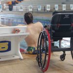 homem de costas sentado na beira da piscina. Atrás dele, está sua cadeira de rodas